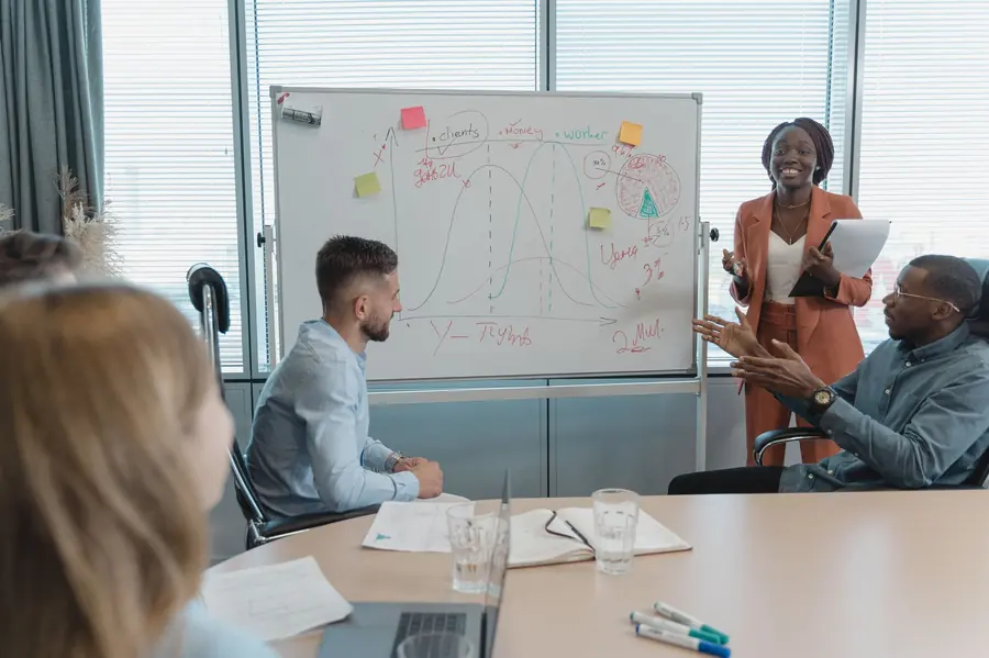 A woman in a rust-coloured suit presenting a whiteboard sketch of two overlapping curves to three seated colleagues in a meeting room